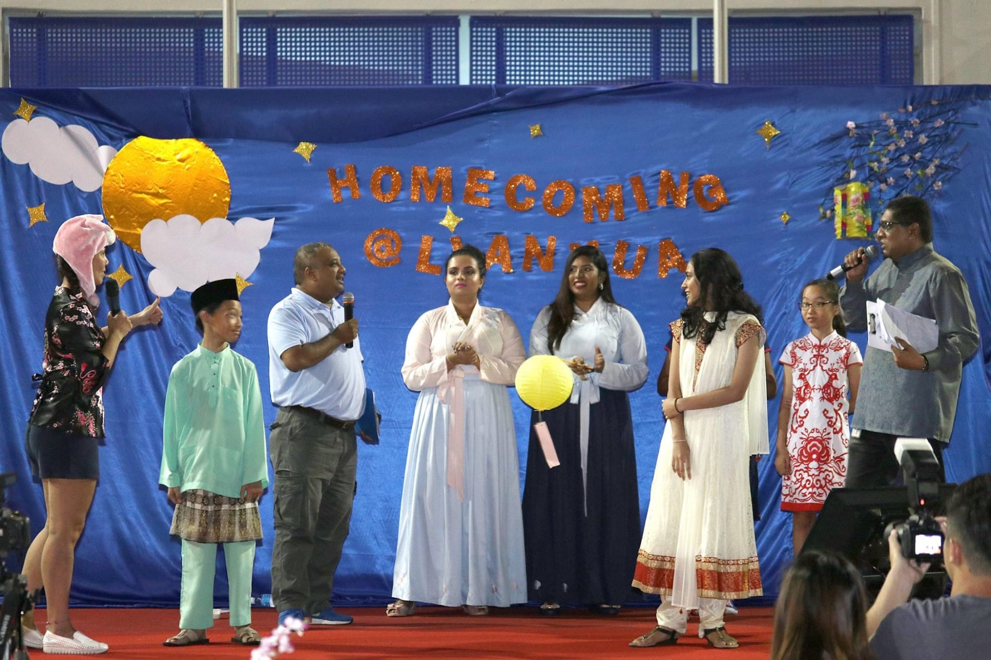 A group of people in traditional outfits stand on a stage before a blue banner reading "Homecoming at Lianmua."